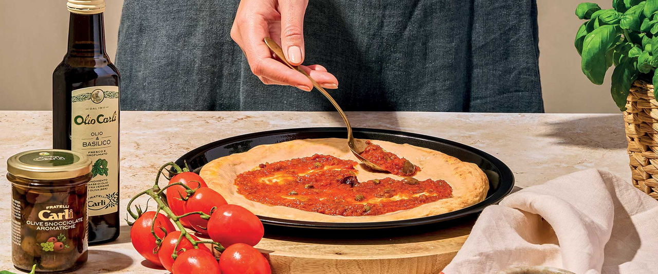 FIGURA FEMMINILE CHE CONDISCE UNA PIZZA CON IL SUGO POMODORO E BASILICO, LE OLIVE SNOCCIOLATE AROMATICHE E L'INFUSO OLIO E BASILICO